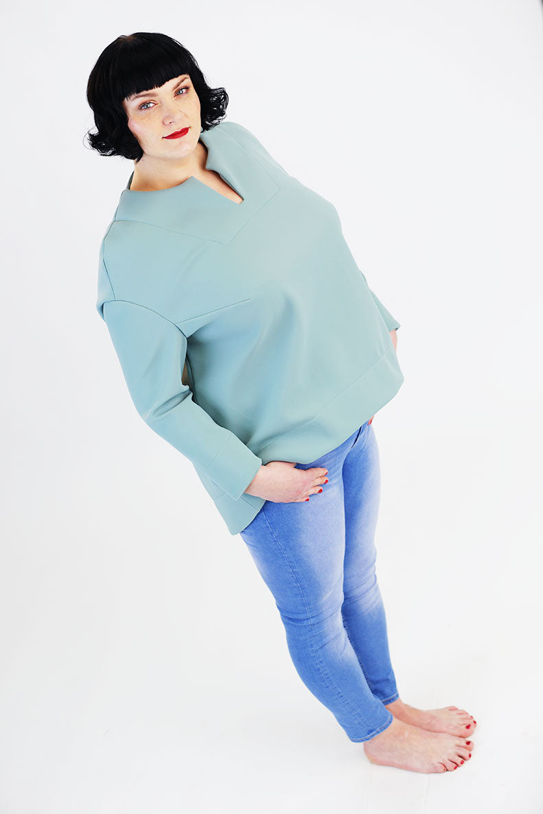 eine Frau steht schräg auf einem weißen Untergrund und trägt ein türkisfarbenes Shirt sowie eine hellblaue Jeans