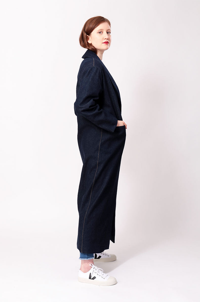 Eine Frau in einem langen Jeansmantel steht vor einem weißen Hintergrund.