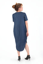 vor einer weißen Wand steht eine Frau in einem blauen Kleid welche in die Ferne schaut