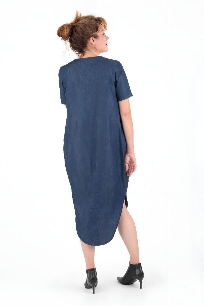 vor einer weißen Wand steht eine Frau in einem blauen Kleid welche in die Ferne schaut