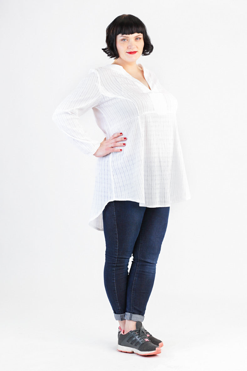 vor einem weißen Hintergrund steht eine Frau mit weißer Tunika und blauer Jeans